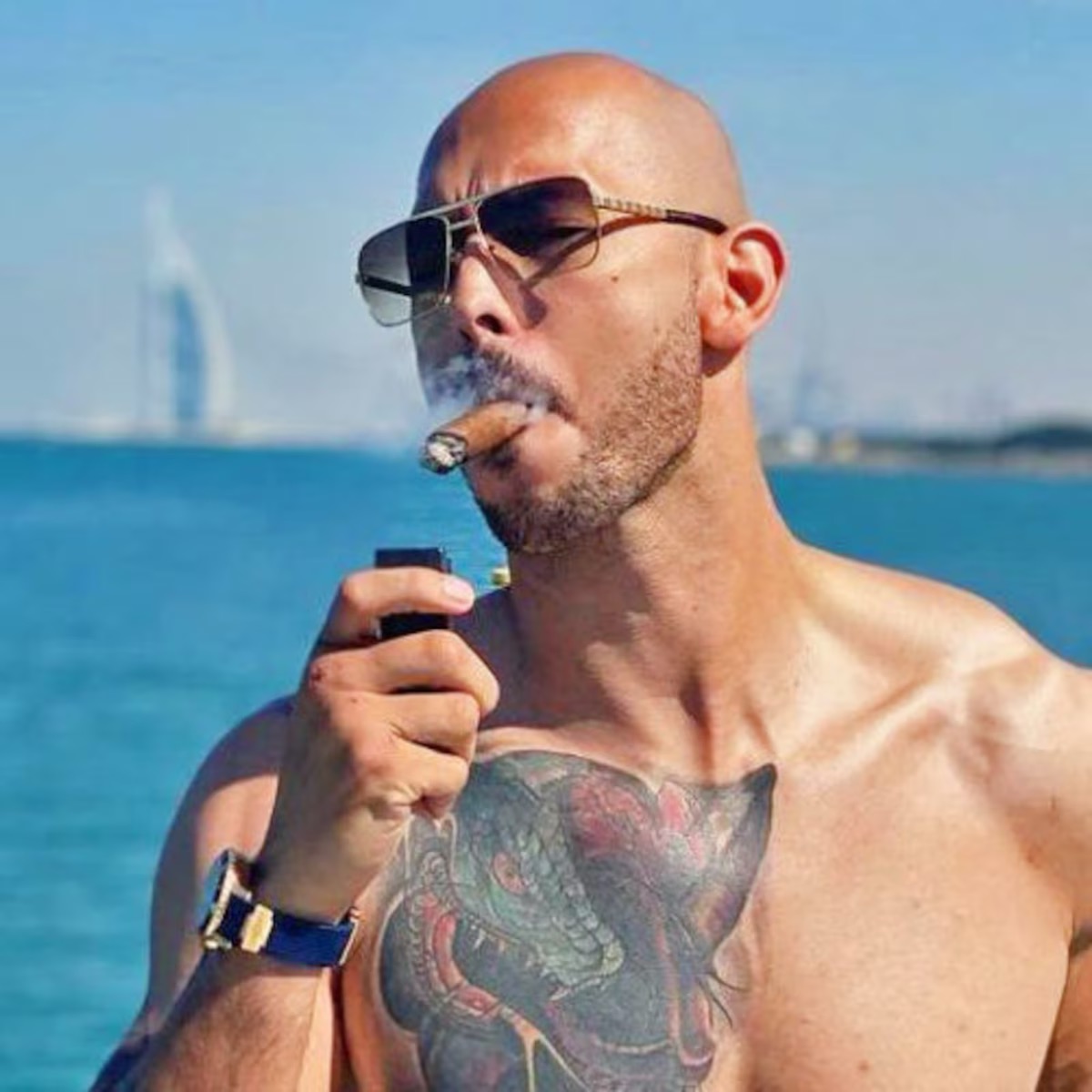 Andrew Tate, shirtless on a Dubai pier with the Burj al-Arab behind him, smoking a cigar.