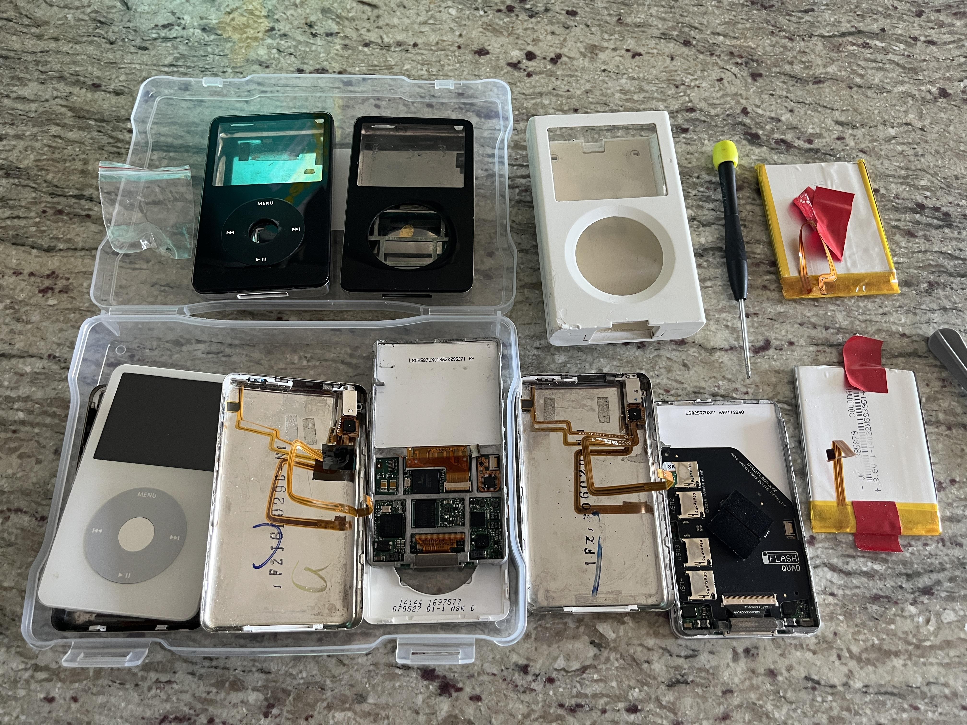 Open iPods on a kitchen counter — replacement batteries with red tape, motherboards, click wheels, screwdriver — mid-modding session.