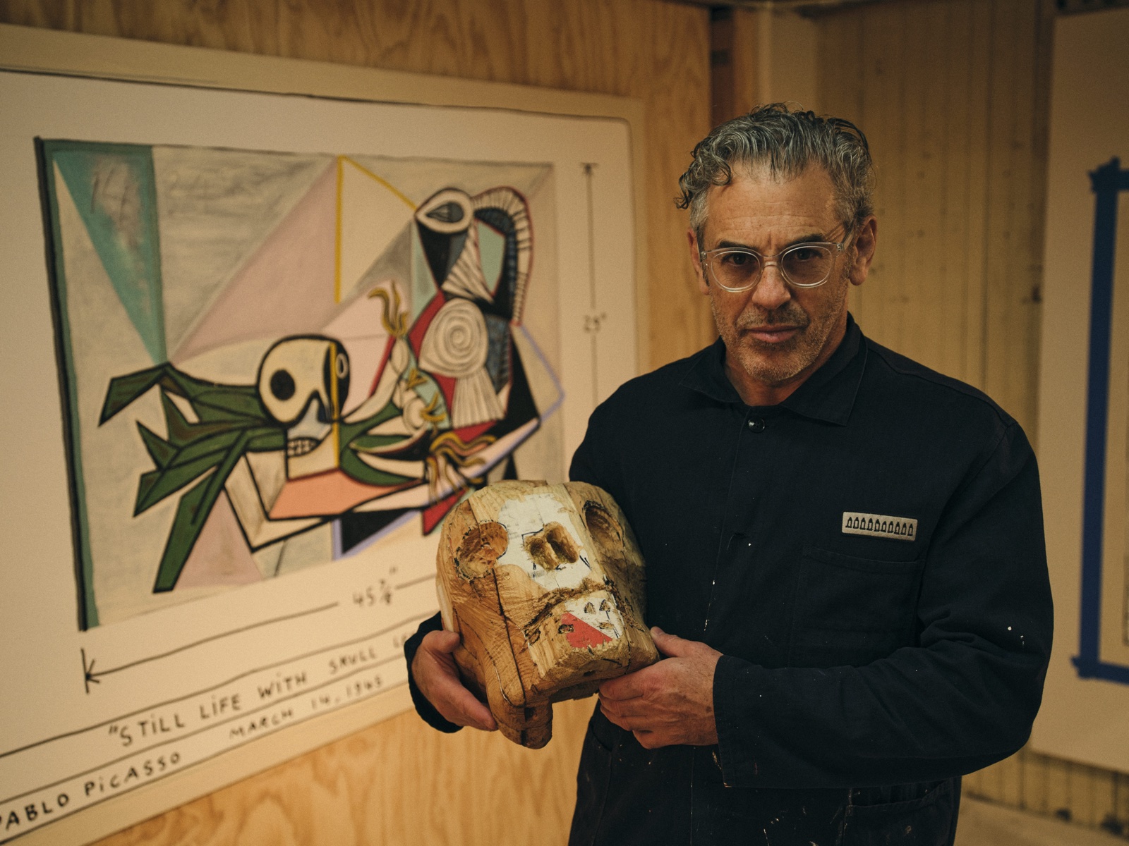 Tom Sachs in his studio, holding a sculpted head in front of a drawing of Picasso's Still Life with Skull.